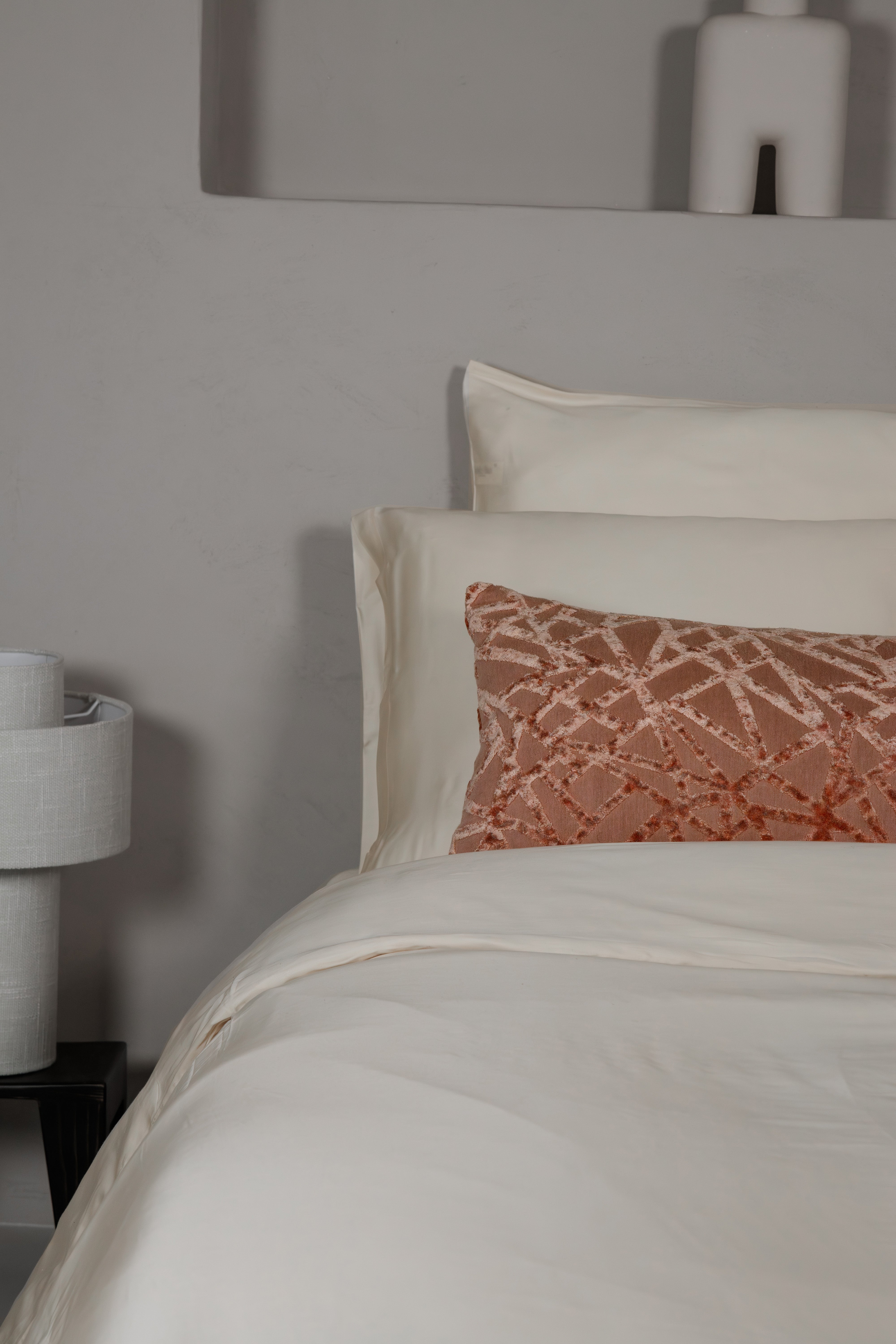 White fitted sheet with matching pillowcases and a textured rust-colored accent pillow on a bed against a grey wall.