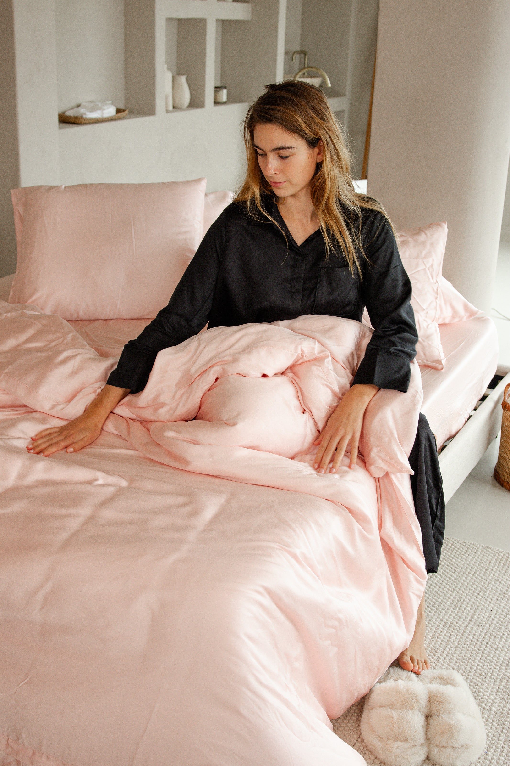 Soft blush pink satin fitted sheet and duvet set on bed with woman arranging duvet.